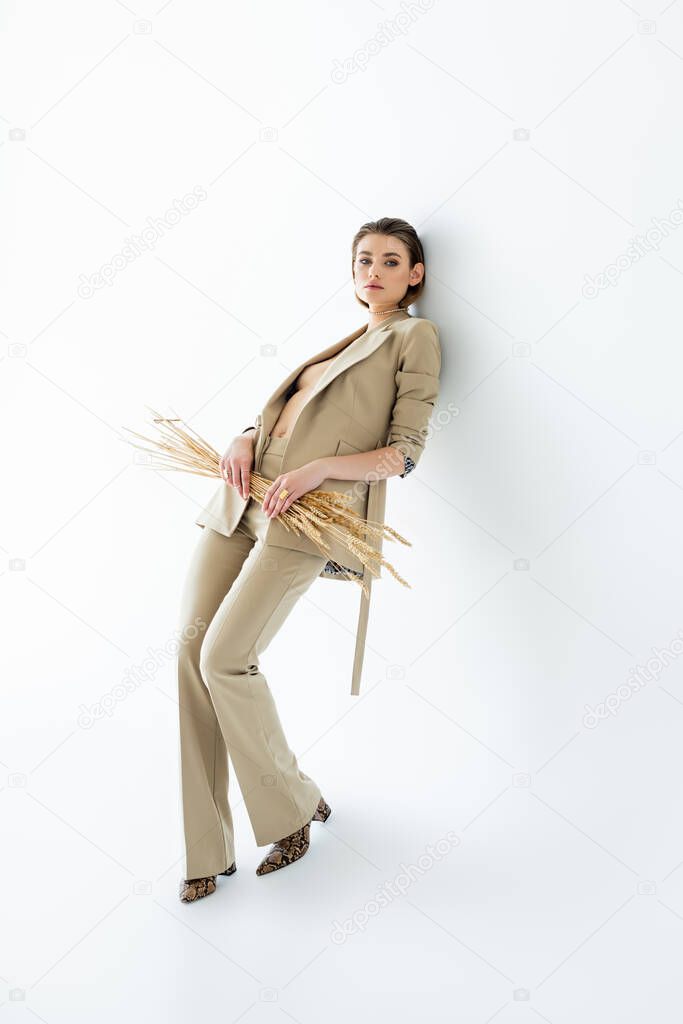 full length of young model in beige suit posing while holding wheat on white