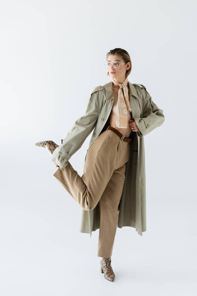 full length of trendy woman in glasses, trench coat and scarf posing on white