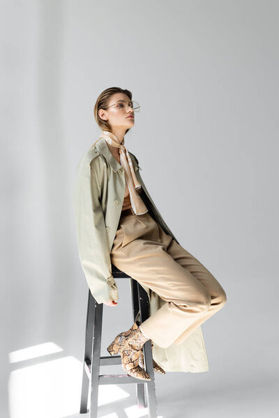 full length of young woman in glasses, trench coat and scarf sitting on stool while posing on grey