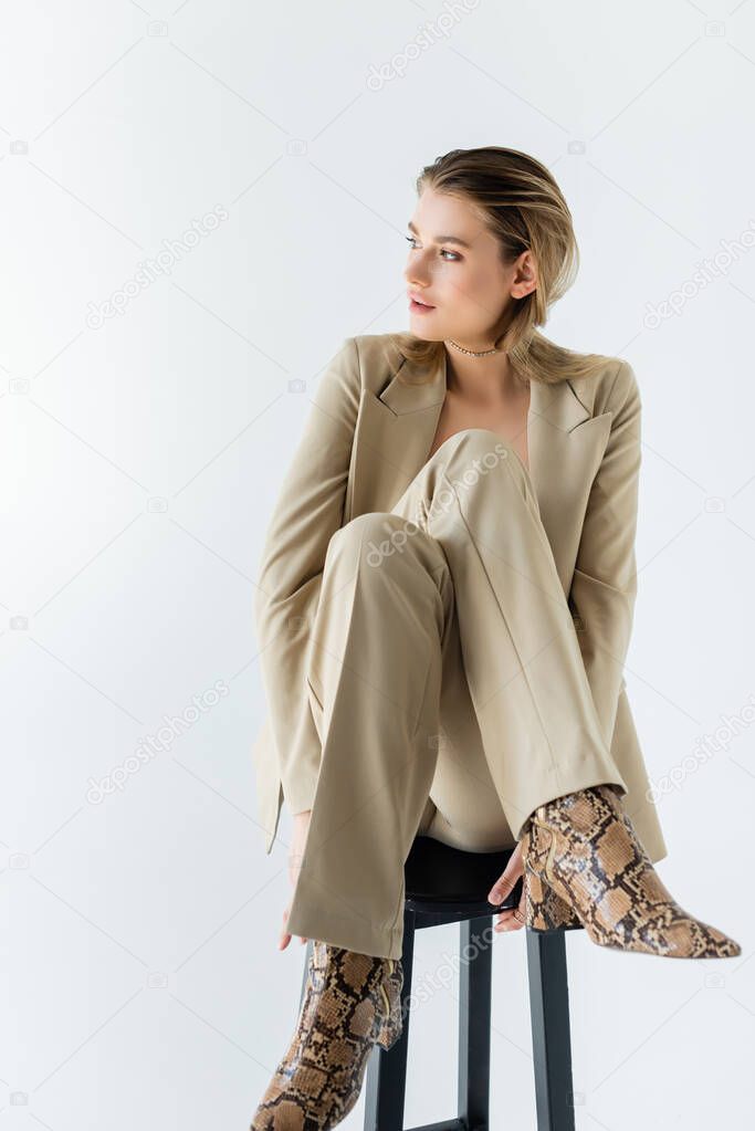 young model in suit sitting on stool and looking away on white background