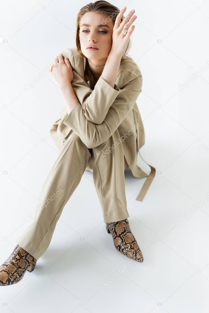 stylish model in beige suit and boots with animal print sitting on white