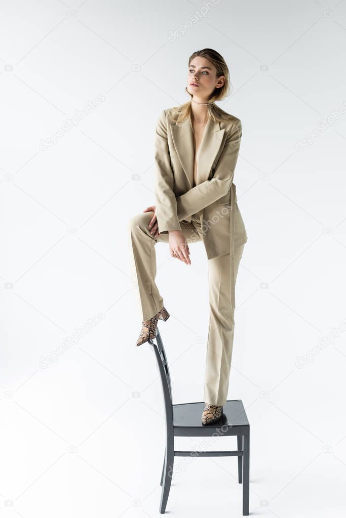 stylish model in beige suit and boots with animal print standing on chair on white