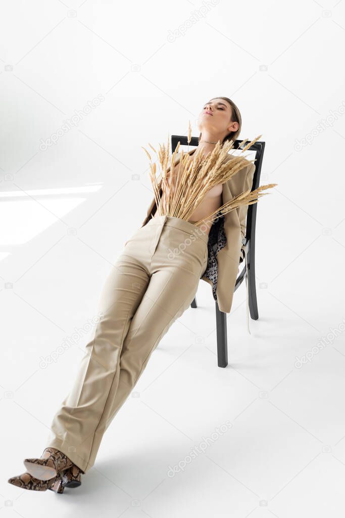 stylish model in beige suit with wheat posing on chair on white background