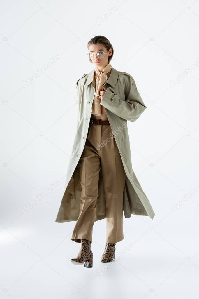 full length of trendy woman in glasses, trench coat and scarf walking on white
