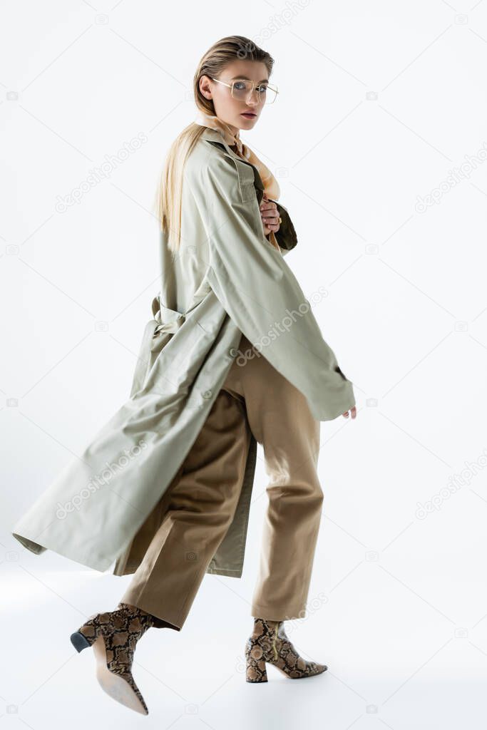 full length of stylish woman in glasses, trench coat and scarf walking on white