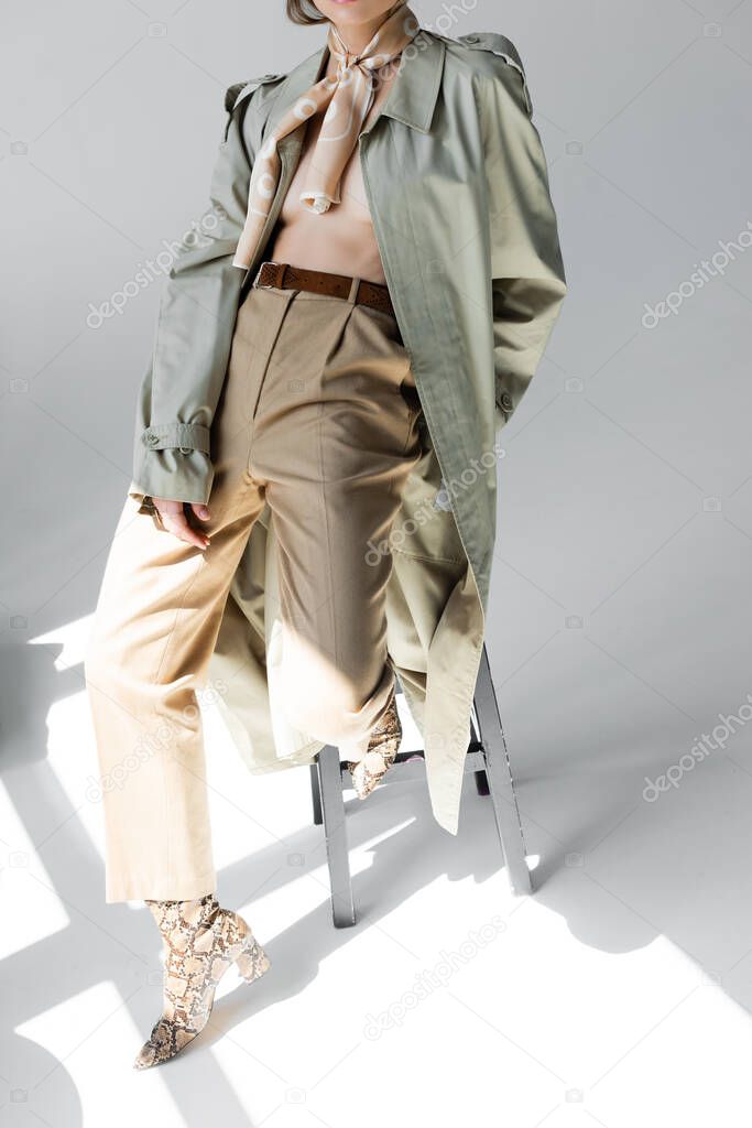 cropped view of young woman in trench coat and scarf leaning on stool while posing on grey