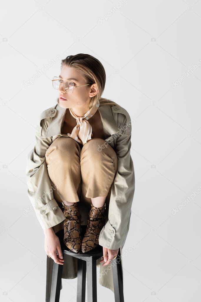 trendy woman in trench coat, glasses and scarf posing on chair isolated on white