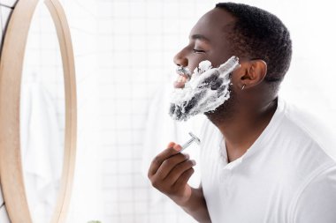 Afro-Amerikan bir adamın tıraş olurken sakalını tıraş edişinin yan görüntüsü. 