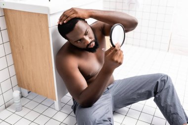 afro-american man sitting on bathroom floor and fixing hair in small mirror clipart