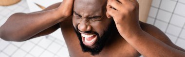 panoramic shot of afro-american man screaming from headache and holding glass of water near temple clipart