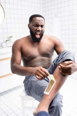 Afro-Amerikalı adam, balmumu şeritlerini yırtıyor ve acıdan bağırıyor. 