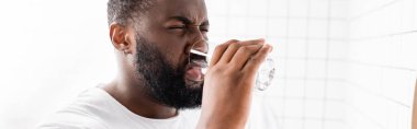 panoramic shot of afro-american man grimacing and drinking water clipart