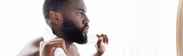 panoramic shot of serious afro-american man looking in mirror with towel behind neck 