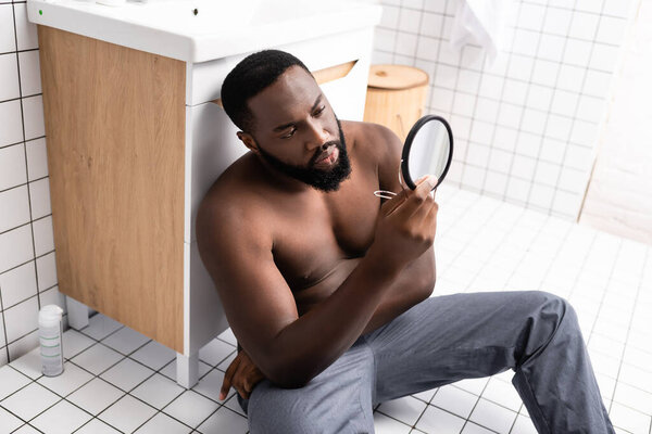 afro-american man sitting on bathroom floor and looking at face in small mirror