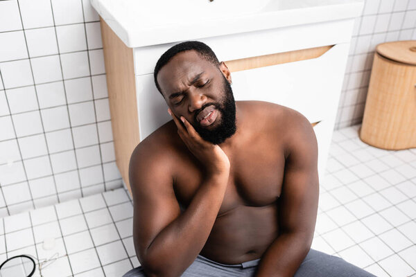 afro-american man sitting on bathroom floor and grimacing from toothache