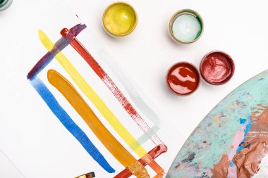 Beyaz arkaplanda fırça darbeleriyle kağıdın yanında Gouache boyaları ve boya fırçasıyla paletin üst görüntüsü