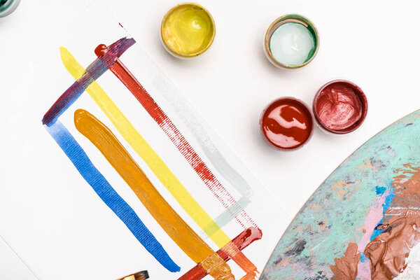 top view of palette with Gouache paints and paintbrush near paper with brushstrokes on white background