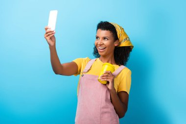 Mutlu Afro-Amerikalı kadın elinde tekrar kullanılabilir kupa ve mavi ile selfie çekiyor.
