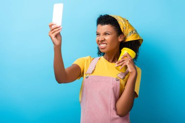 Kıvırcık Afrikalı Amerikalı kadın tekrar kullanılabilir kupa tutuyor ve mavi fincanda selfie çekerken dişlerini gösteriyor.
