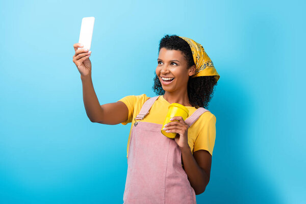 happy african american woman holding reusable mug and taking selfie on blue