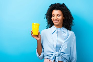 happy african american woman holding empty reusable mug on blue clipart