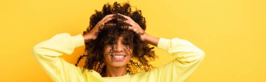 happy african american woman with closed eyes fixing curly hair on yellow, banner clipart
