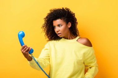 Şok olmuş Afrikalı Amerikalı kadın sarıdaki klasik telefona bakıyor.