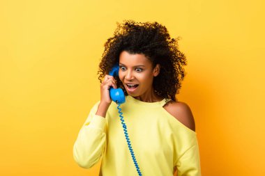 Şok olmuş Afro-Amerikan kadın sarıda klasik telefondan konuşuyor.