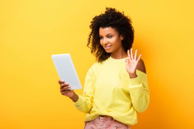 Afro-Amerikalı kadın dijital tablet tutarken ve el sallarken sarıda görüntülü sohbet ederken