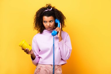 Şok olmuş Afrikalı Amerikalı kadın mavi retro telefondan konuşuyor ve elinde sarı bardakla eko bardağı tutuyor.