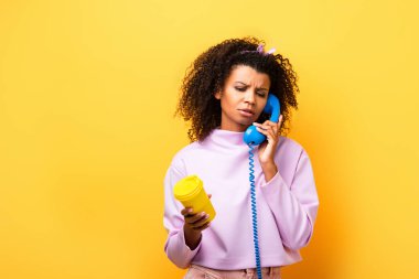 Afrikalı Amerikalı kadın mavi retro telefondan konuşuyor ve sarı bardakta eko bardağı tutuyor.