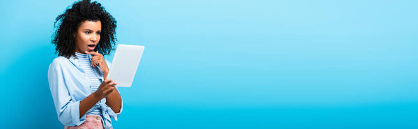 shocked african american woman using digital tablet on blue, banner