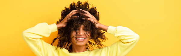 happy african american woman with closed eyes fixing curly hair on yellow, banner