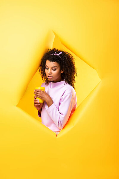 african american woman holding eco cup and looking down through hole in ripped paper on yellow background 