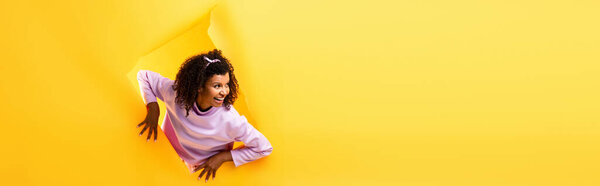 curious african american woman looking away through hole in ripped paper and smiling on yellow background, banner
