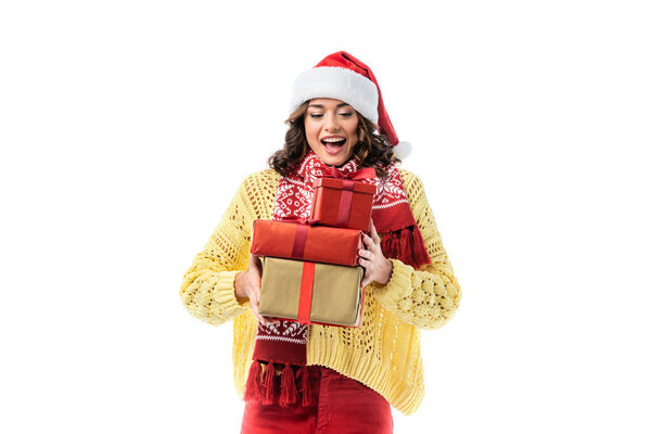 joyful young woman with open mouth, in santa hat and scarf holding gifts isolated on white