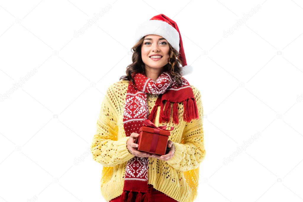 young pleased woman in santa hat and red scarf holding present isolated on white
