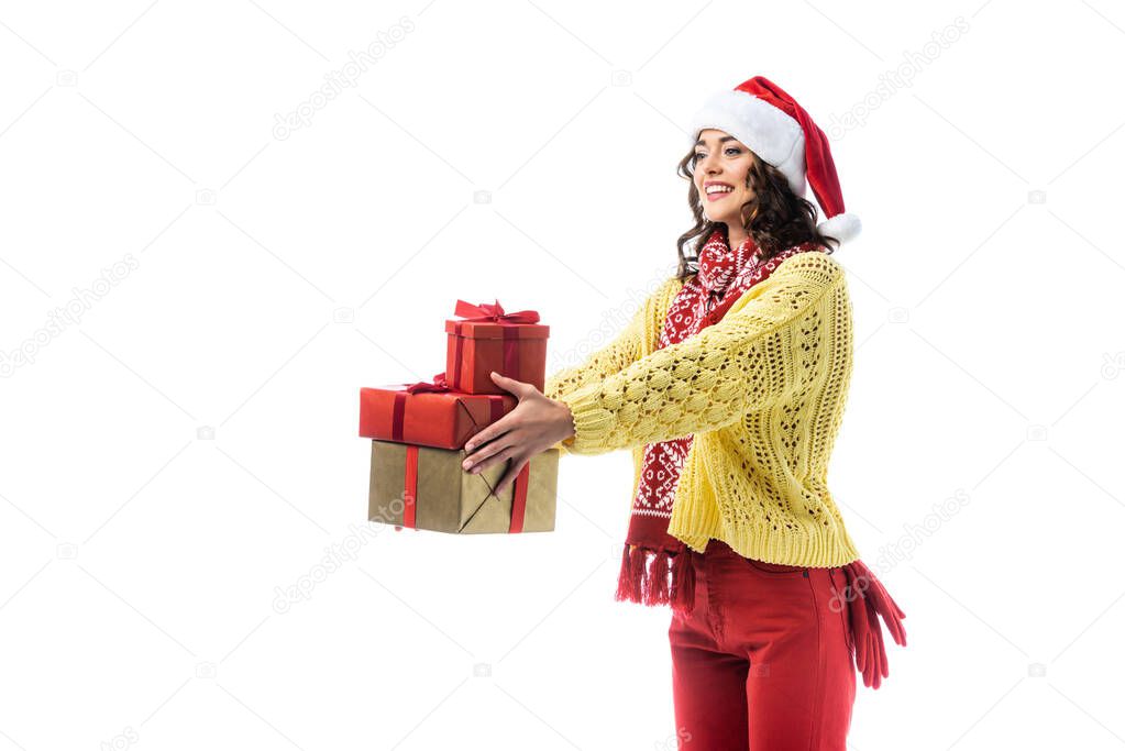 joyful young woman in santa hat, scarf and knitted sweater holding gifts isolated on white