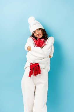 young woman in winter outfit, warm scarf, gloves and hat embracing herself on blue clipart