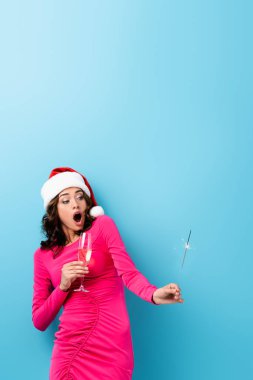 Şok olmuş Noel Baba şapkalı kadın elinde maytap ve mavi üzerine bir bardak şampanya tutuyor.