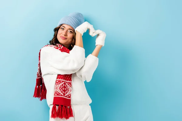 pleased young woman in winter outfit, warm scarf, gloves and hat showing heart sign on blue