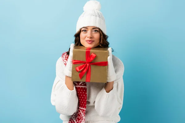 joyful woman in winter outfit holding wrapped present and looking at camera on blue