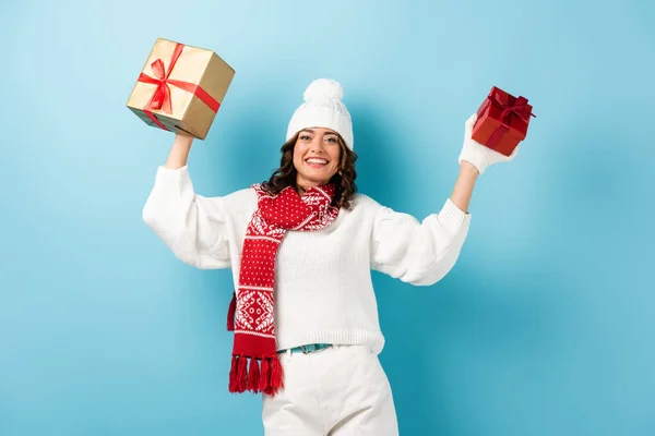 young pleased woman in winter outfit holding presents on blue