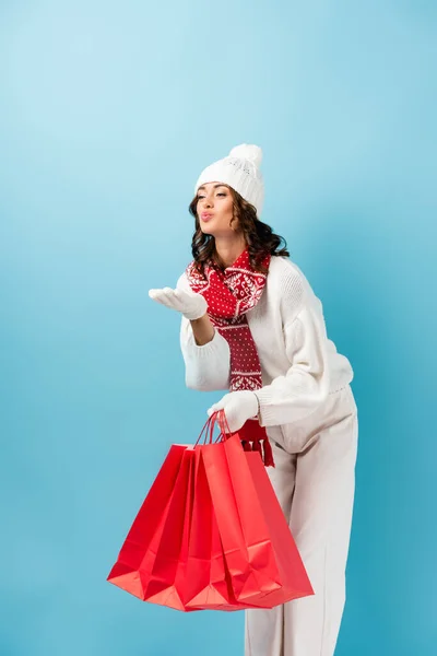 young woman in winter outfit holding red shopping bags and sending air kiss on blue