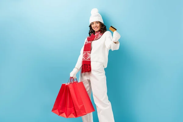 happy young woman in winter outfit holding red shopping bags and credit card on blue