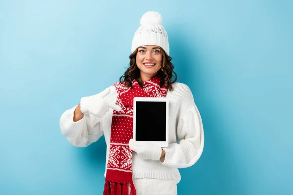 joyful woman in winter outfit pointing with finger at digital tablet with blank screen on blue