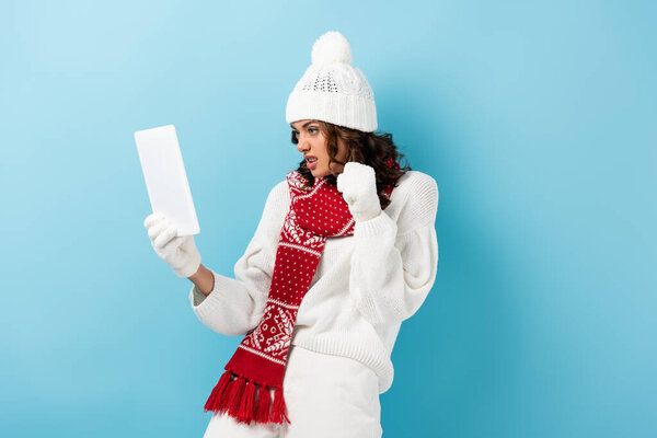 angry young woman in winter outfit looking at digital tablet on blue