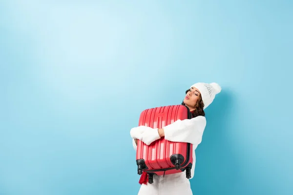 young woman in winter outfit carrying heavy suitcase on blue