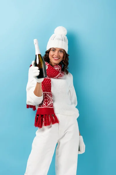 joyful woman in winter outfit holding bottle of champagne on blue