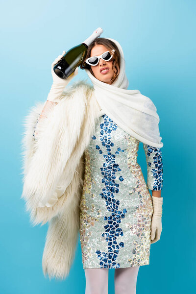 displeased woman in sunglasses holding bottle of champagne on blue